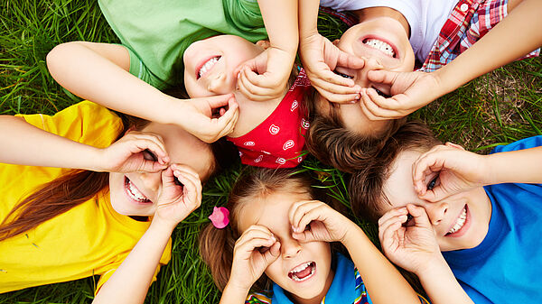 Mehrere Kinder liegen mit den Köpfen im Kreis auf dem Boden und schauen durch kreisförmige "Lupen", die sie mit Daumen und Zeigefinger zu Ringen geformt haben