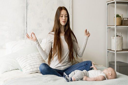 Junge Frau macht eine Yoga-Pose im Bett und das Baby liegt neben ihr und schaut sie an.
