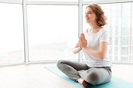 Junge Frau in einer Yoga-Pose sitzt auf der Matte in einem Raum mit gro&szlig;en Fenstern.
